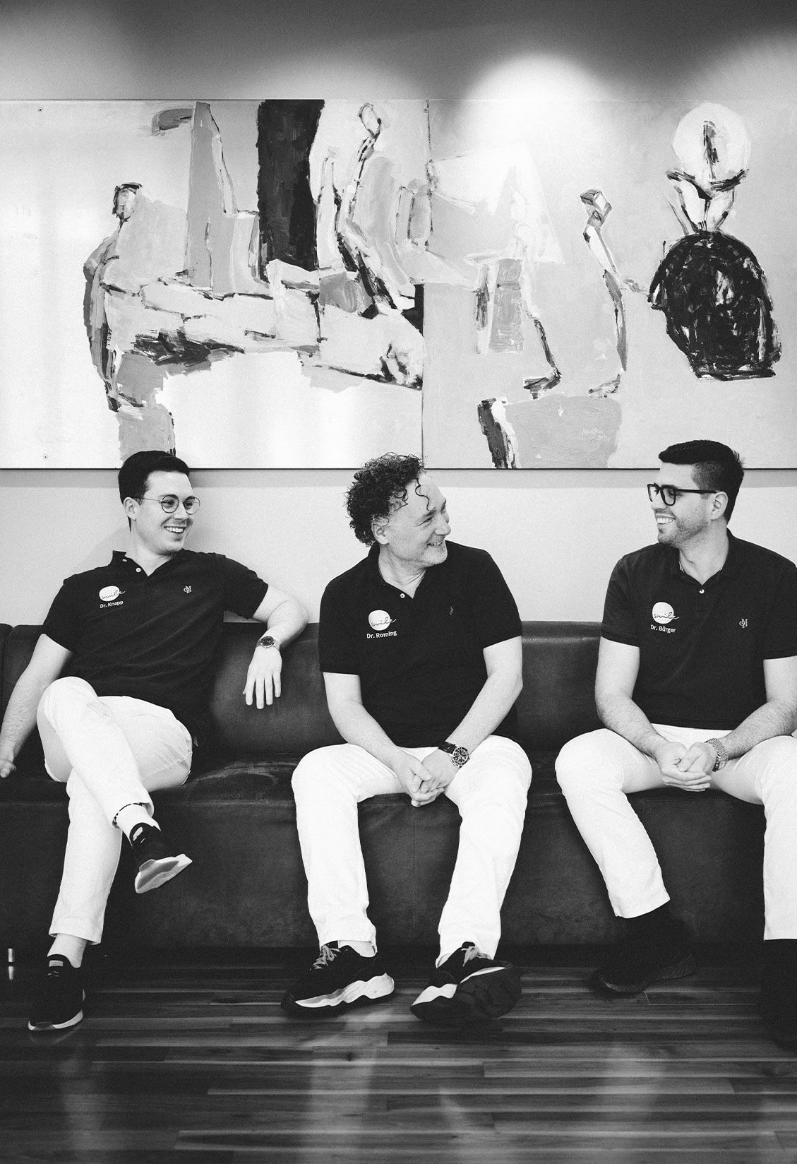Kieferorthopädie Deggendorf – Dr. Roming, Dr. Knapp und Dr. Bürger sitzen gemeinsam auf einem Sofa, lachen einander zu und tragen dunkle Poloshirts mit Namensstickerei; im Hintergrund ein abstraktes Gemälde, das Bild ist in Schwarz-Weiß gehalten und vermittelt eine herzliche, kollegiale Atmosphäre.