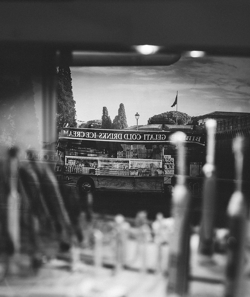 Kieferorthopädie Deggendorf – Schwarz-Weiß-Fotografie eines Wandmotivs mit Spiegelung: Ein nostalgischer Eiswagen mit der Aufschrift „Gelati Cold Drinks Ice Cream“ vor klassischer Stadtkulisse, im Vordergrund verschwommen zahnmedizinische Instrumente – künstlerisch eingefangene Verbindung von Praxisalltag und urbaner Ästhetik.