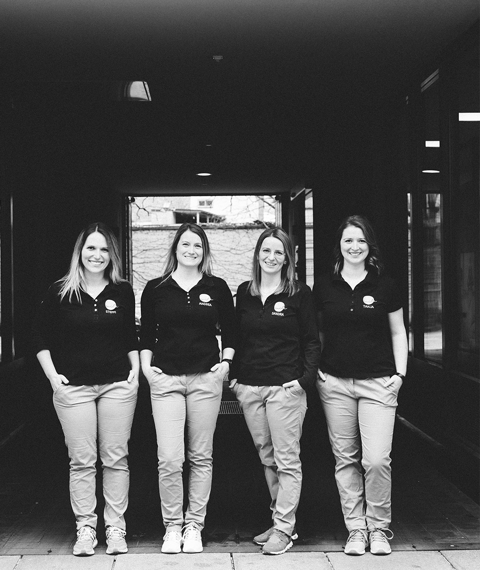 Kieferorthopädie Deggendorf – Vier Mitarbeiterinnen von ARTPRAXIS® stehen lächelnd nebeneinander in einem überdachten Durchgang; sie tragen einheitliche Teamkleidung – das Schwarz-Weiß-Foto vermittelt Teamgeist und professionelle Ausstrahlung.